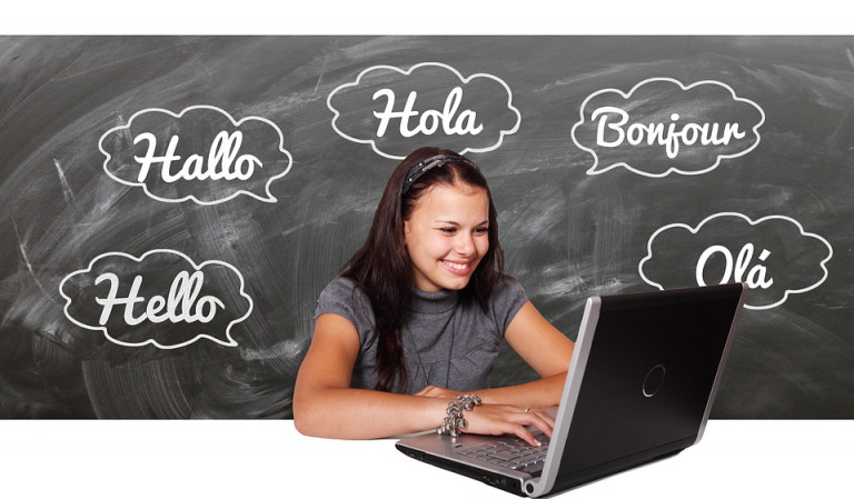Dziewczynka z komputerem, laptopem. W tle tablica z napisem dzień dobry w obcych językach