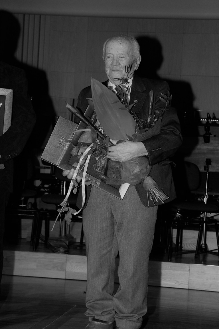 Czarno biała fotografia. Świętej pamięci Henryk Dłużewski stoi na scenie, trzymając w rękach wyróżnienia i bukiet kwiatów