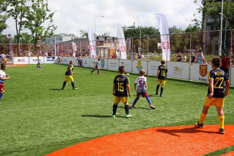 Turniej piłkarski Piątki na Rynku - Sport CK. Na małym boisku, ogrodzonym siatką, młodzi chłopcy, ubrani w stroje sportowe, rozgrywają mecz piłki nożnej.