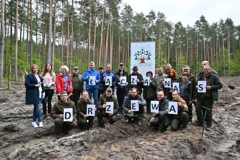 Uczestnicy akcji pozują do pamiątkowego zdjęcia po zakończeniu sadzenia młodych drzewek. W rękach trzymają kartki papieru z literami, nazwą akcji, Łączą Nas Drzewa