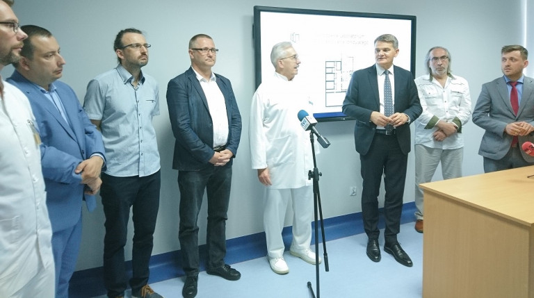 Mężczyźni w garniturach, lekarze w białych fartuchach, stoją przed mikrofonami. Za nimi, na ścianie telewizor. Napis samodzielne laboratorium promieniowania jonizującego