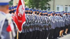 Funkcjonariusze w mundurach galowych stoją na baczność na placu. W tle rozmyty budynek komendy. Na pierwszym planie czerwony sztandar Policji