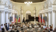 Koncert pianisty zdjęcie z daleka przedstawiające także publiczność. Na scenie w sanatorium Marconi mężczyzna ubrany na czarno gra na fortepianie. Przed i za fortepianem flagi państw