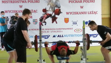 Sportowiec leżąc próbuje podnieść ciężką sztangę. Po bokach asystenci czuwający nad bezpieczeństwem zawodnika mistrzostw Polski w wyciskaniu leżąc