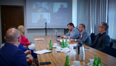 Sala konferencyjna, uczestnicy spotkania siedzą przy stole. Na ścianie obraz z rzutnika ekranu, miniaturki uczestniczących zdalnie