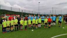 Na boisku piłkarskim do zdjęcia pozują dwie drużyny piłkarskie. W pierwszym rzędzie młode dzieci. W drugim, drużyna dorosłych zawodników futbolu