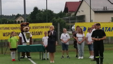 Wicemarszałek Renata Janik z mikrofonem przemawia do zgromadzonych na boisku piłkarskim uczestników wielkiego turnieju charytatywnego. Obok dziewczynki w stroju piłkarskim stoi duża maskotka misia