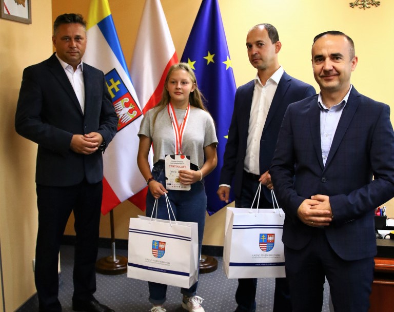 Na tle flag województwa, Polski i Unii Europejskiej, pozują do pamiątkowej fotografii członek zarządu województwa Tomasz Jamka, kielczanka wicemistrzyni Europy w Sumo z trenerem, oraz dyrektor Piotr Kisiel