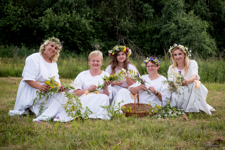 Grupa kobiet w białych sukienkach siedzi na trawie. Na głowach mają wianki z kwiatów