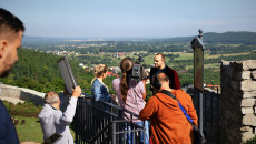 Grupa osób na terenie zamku w Chęcinach. Ekipa telewizyjna z kamerą, mikrofonem i planszą odbijającą światło słoneczne realizuje program na żywo