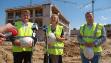 Trzy dźwigi na placu budowy urzędu miar w Kielcach. Trzy osoby stoją na tle budynku w stanie surowym. Na pytania dziennikarzy odpowiada wicemarszałek Renata Janik