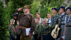 Mężczyzna ubrany w historyczny mundur salutuje. Obok grupa harcerzy i zuchów. Radny Województwa Paweł Krakowiak prowadzi apel poległych