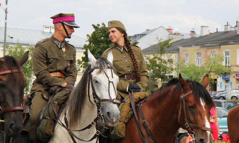 Młodzi kobieta z warkoczem i mężczyzna ubrani historyczne wojskowe mundury jadą konno. Uczestnicy Kadrówki na ulicach miasta Kielce