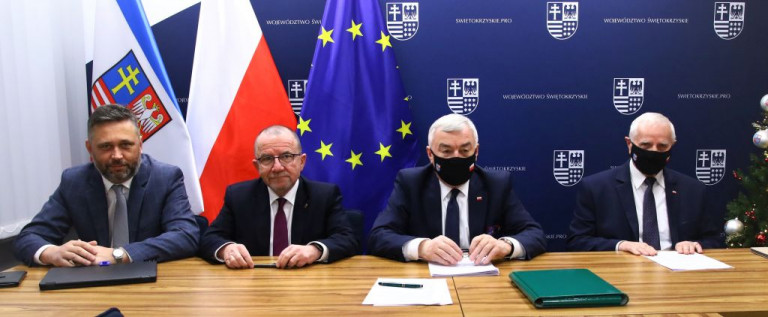 Czterej mężczyźni w garniturach siedzą przy stole, na którym leżą dokumenty. Za mężczyznami flagi, od lewej: Województwa Świętokrzyskiego, Polski i Unii Europejskiej