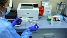 Przy biurku na którym ustawione jest urządzenie do przeprowadzania badań siedzi laborantka z fiolkami w dłoniach