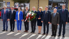 Uczestnicy uroczystości stoją z wiązanką kwiatów na ulicy Warszawskiej, naprzeciw pomnika P. Gosiewskiego