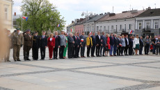 Uczestnicy uroczystości stoją przed masztem, na który wciągana jest flaga państwowa