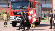 Strażacy maszeruja ze sztandarem na tle nowego wozu strażackiego