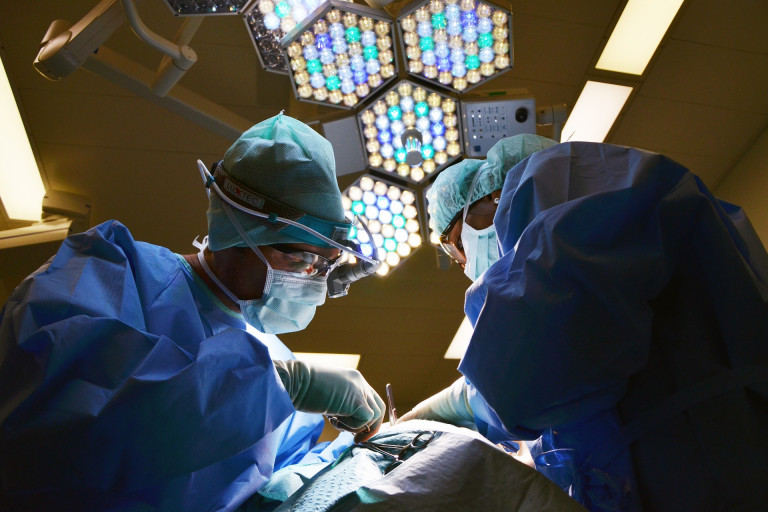 2 Chirurdzy w Maskach I Sprzęcie Pochyleni Nad Pacjentem Na Sali Operacyjnej, W Tle Lampa Chirurgiczna
