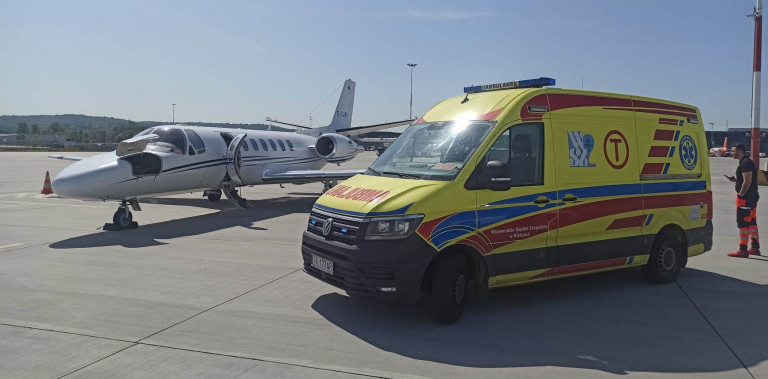 Na lotnisku biały mały samolot, przed samolotem żółty pojazd transportu medycznego. Obok karetki stoi ratownik