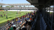 Publiczność widowiska siedzi na trybunie stadionu, w tle zawodnicy rozgrywają mecz na stadionie